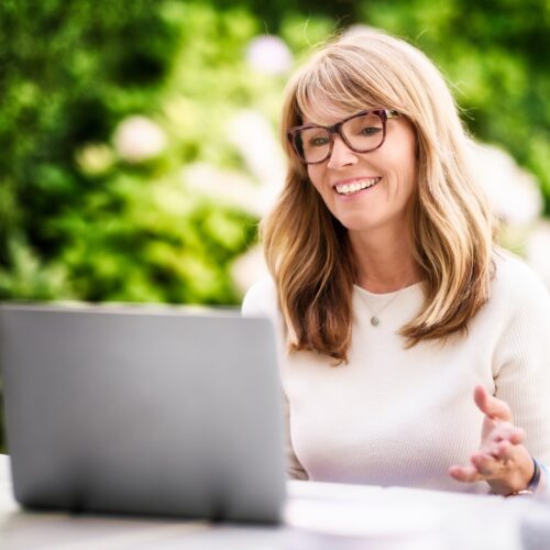 Happy mature woman sitting at home in the garden and using laptop while having video call.