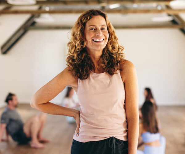 Mature woman standing in a fitness studio Mature woman smiling at the camera while standing in a fitness studio with a group of people in the background. Happy woman having an exercise session with her class in a community yoga studio.