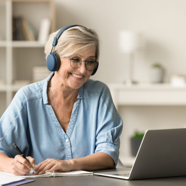 Happy senior retired student woman in earphones and glasses studying on Internet from home, writing notes at laptop, watching online educating lecture, smiling, laughing