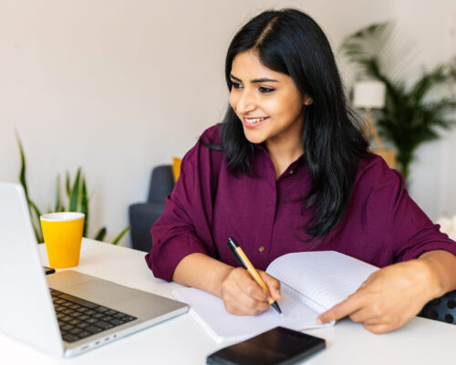 Young adult indian student woman taking notes while using laptop at home Young adult indian student woman taking notes while using laptop computer at home. Millennial ethnic female learning online listening virtual video call. Business and education concept.