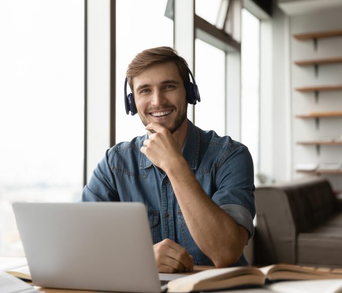 Portrait of happy millennial generation male student. Portrait of happy millennial generation man in headphones sitting at table with books and computer. Smiling young male student posing in modern home office, e-learning distantly on online courses.