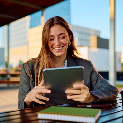 Professional young businesswoman smiling while working with a digital tablet on an outdoor terrace, showcasing modern business, technology, and career success in a relaxed environment Professional woman smiling using tablet outdoors