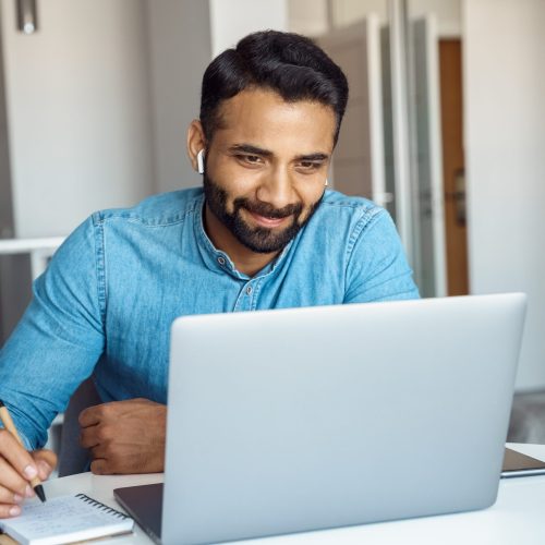 Portrait of young adult indian man wearing earphones looking at laptop screen Portrait of young adult indian man wearing earphones looking at laptop screen. Motivated student writing notes during online lesson, watching webinar, learning language online, sitting at work desk